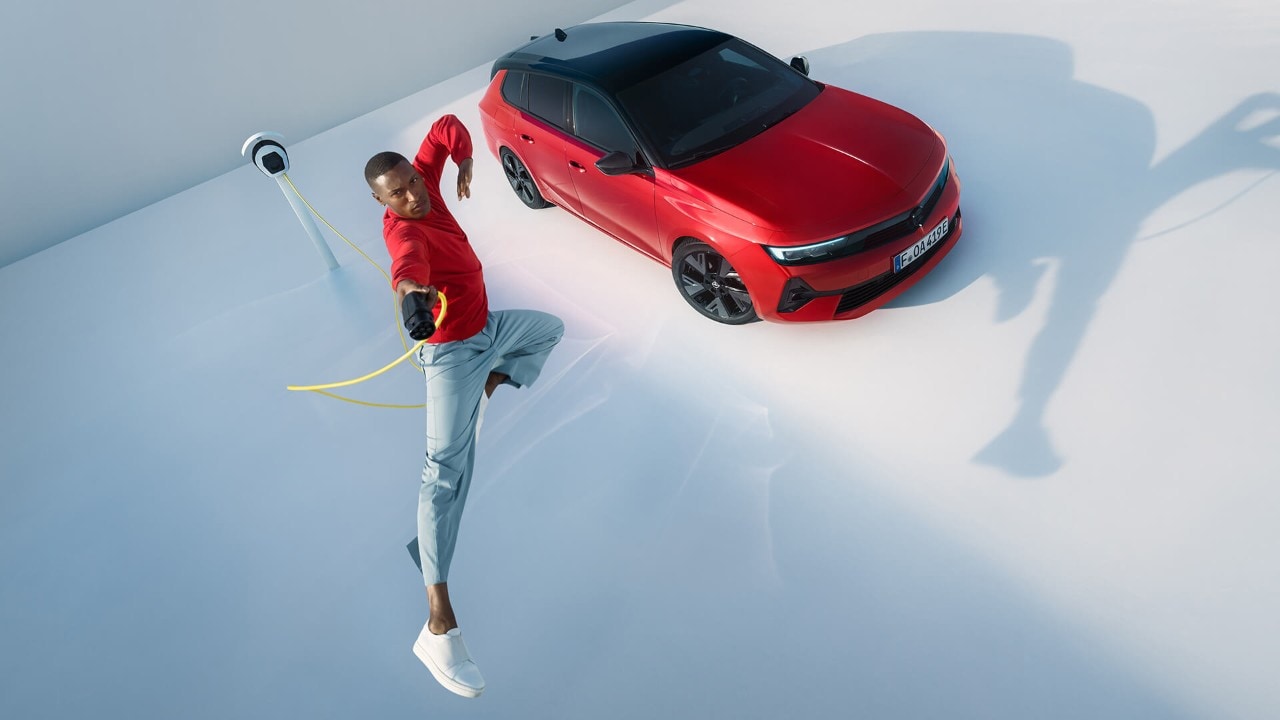 Ein Mann springt in die Luft, während im Hintergrund eine Ladestation für Elektrofahrzeuge und ein Opel Astra Sports Tourer Electric zu sehen sind.