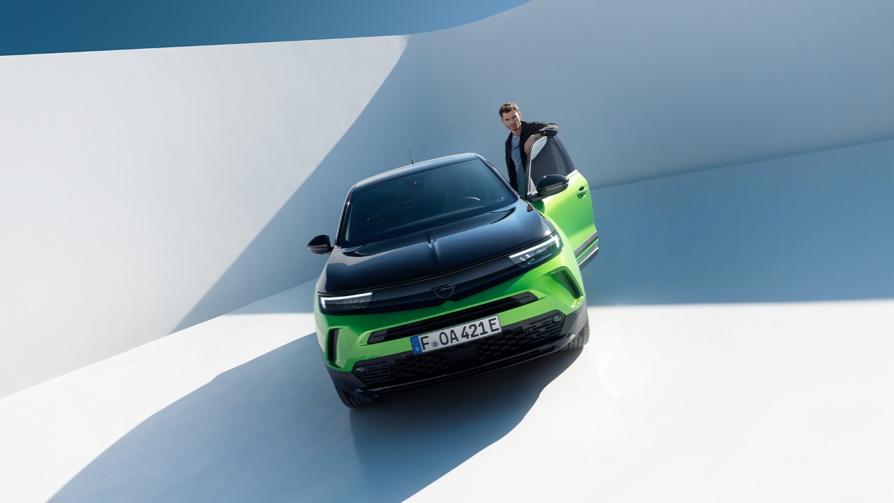Frontansicht eines grünen Opel Mokka Electric mit schwarzen Felgen und Dach mit einem Mann, der über die Fahrertür einsteigt